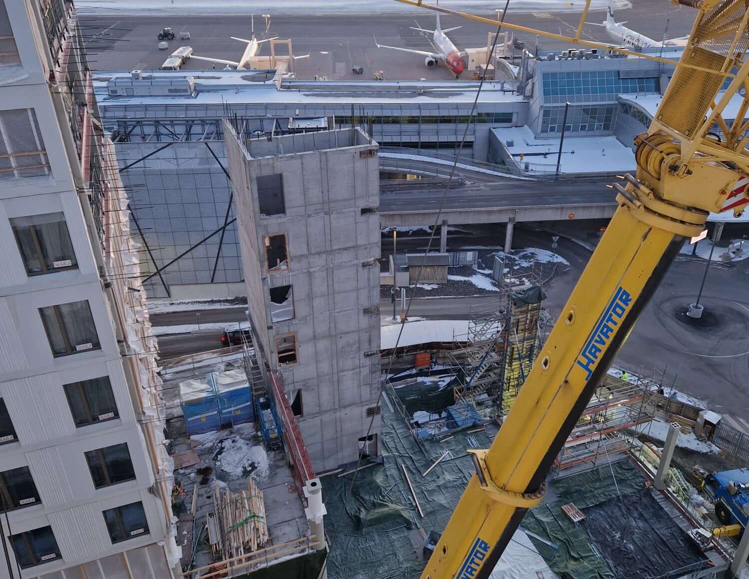 Rakennustyömaa lentoaseman vieressä. Keltainen nosturi avustaa betonielementtien asennuksessa. Kuvassa näkyy keskeneräinen hissikuilu ja ympärillä hotellirakenteita.