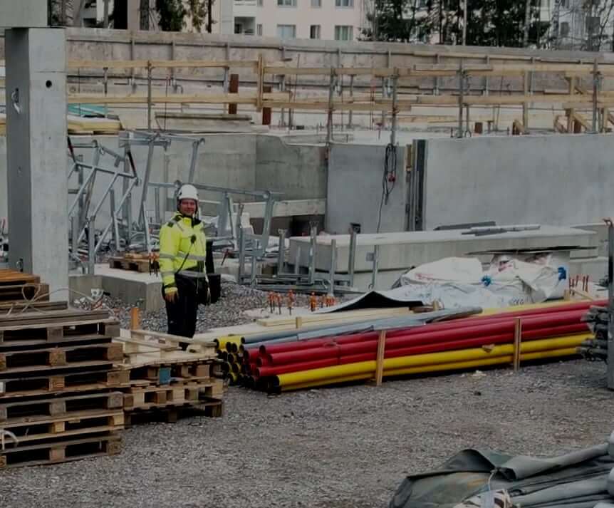 Rakennustyömaa, jossa työntekijä seisoo betonielementtirakenteiden keskellä. Maassa on varastoituna värikoodattuja putkia, kuormalavoja ja rakennusmateriaaleja.